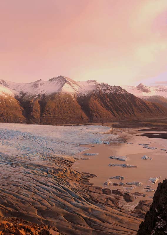 Large glacier flowing into a waterway, surrounded by mountains, viewed from above at sunset