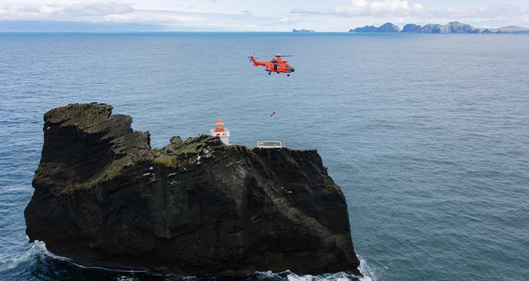 A helicopter hovers a small island with a lighthouse on it.